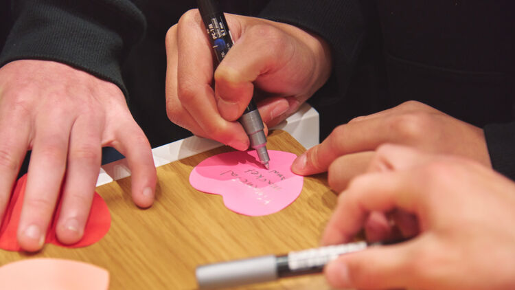 A close-up image of hands using a pen to write a message on a pink heart-shaped card.