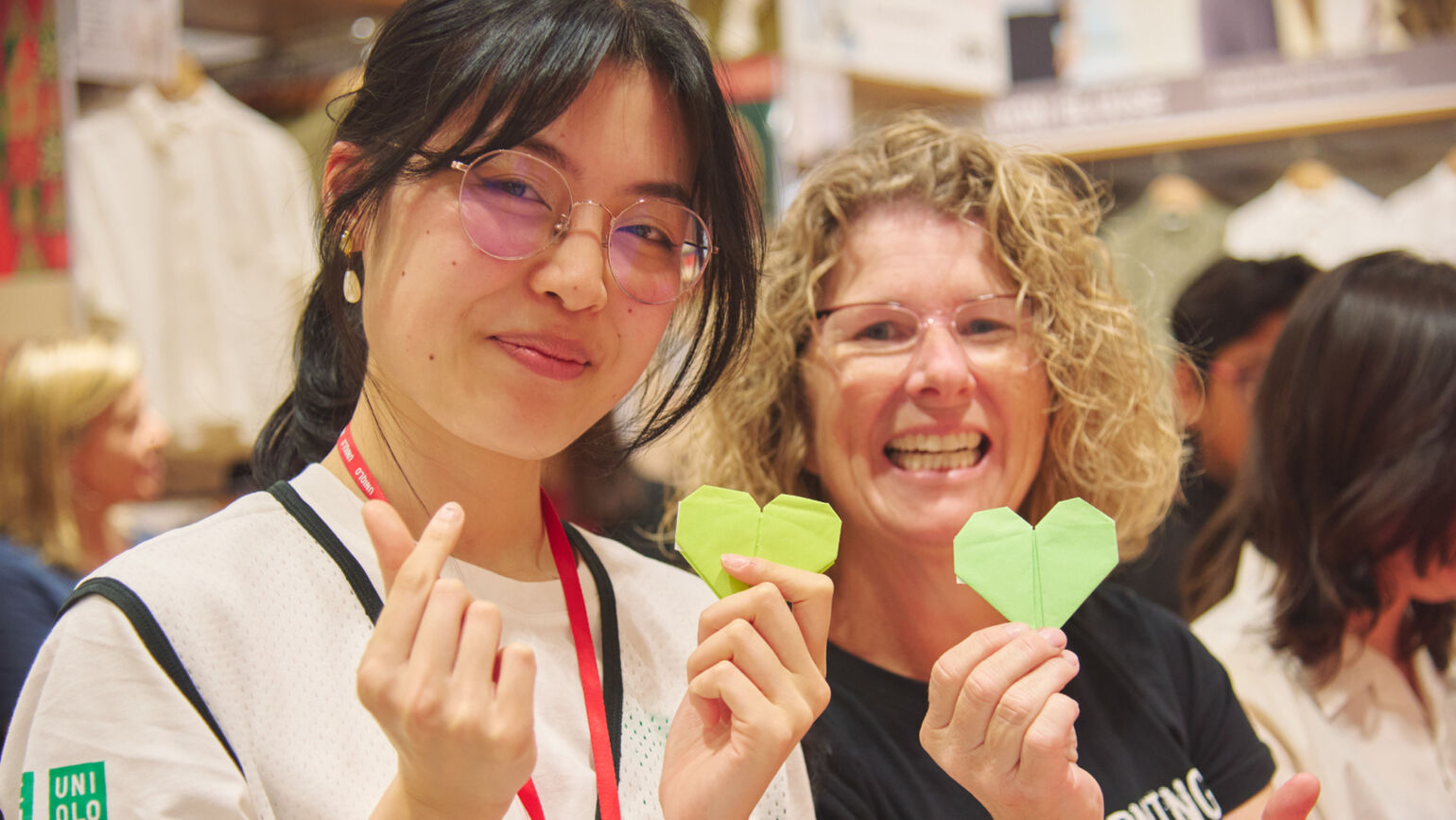 An image of a woman wearing glasses and another smiling woman standing together, each holding a newly folded green origami heart.