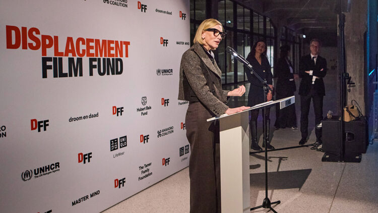 An image of Cate Blanchett standing at a podium and giving a speech on a stage with activity logos lined up in the background.
