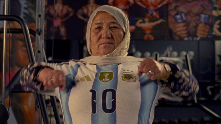 An image of an elderly woman wearing an Argentina national football team jersey, with a powerful expression in front of gym training equipment.