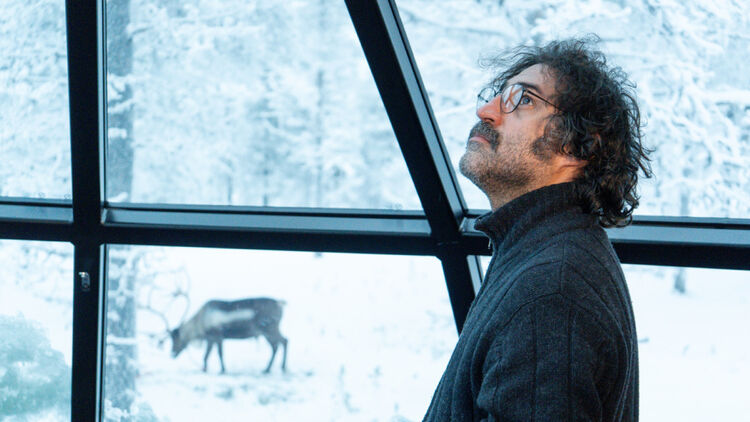 An image of a man wearing glasses looking up from inside a room at a snowy landscape outside the window.
