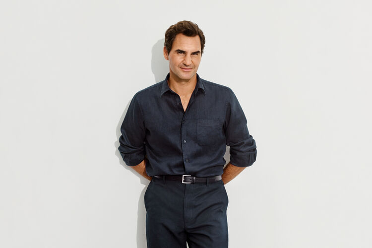An image of Roger Federer looking smartly dressed in a dark navy linen shirt, standing with his hands on his hips and gazing forward.