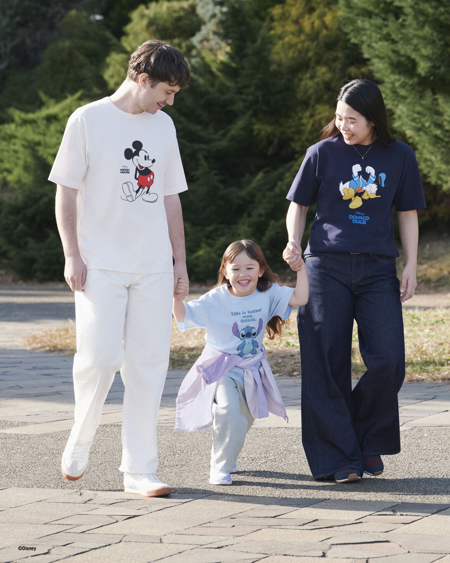 An image of the Hobbs Family—Jaimy, Daisy, and Seira—smiling and walking hand-in-hand under the sunlight, wearing T-shirts featuring Mickey Mouse, Donald Duck, and Stitch.