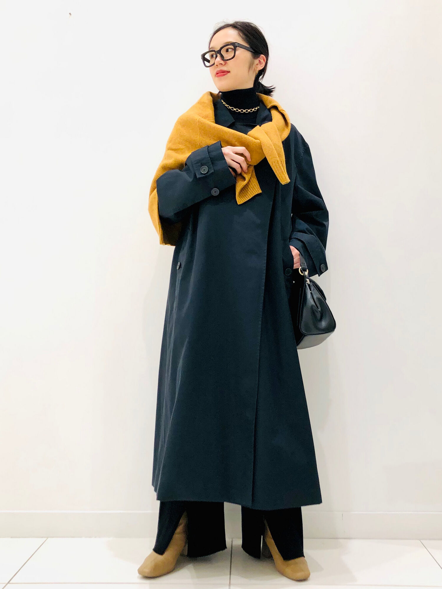 明るいイエローのセーターを肩がけした、トレンチコートを使ったオールブラックのレディースコーデ。