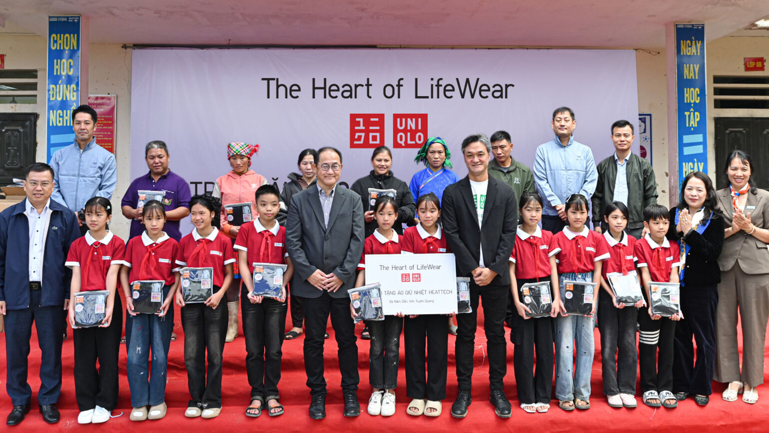 ユニクロの横断幕の前で、子供たちと関係者がヒートテックを手に並ぶ集合写真の画像