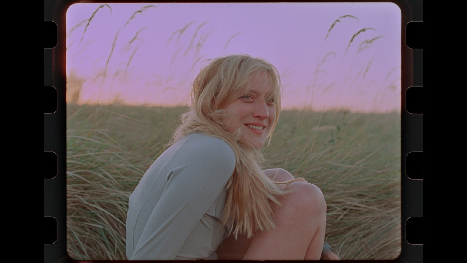 夕暮れ時の草原で、金髪の女性がこちらを向いて穏やかに微笑んでいる画像。