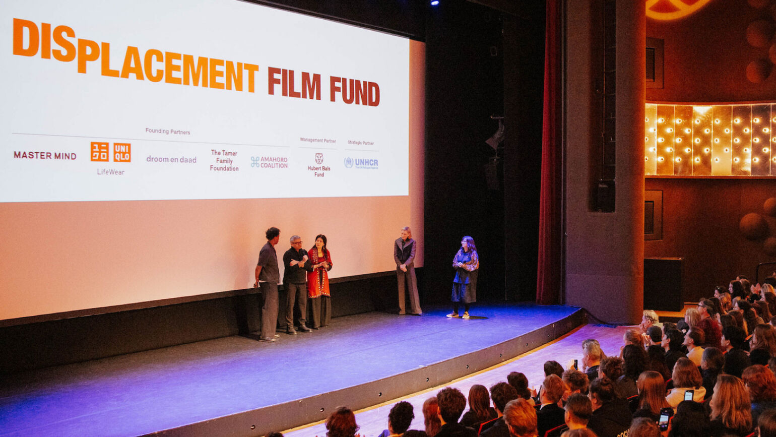 「DISPLACEMENT FILM FUND」と大写しになったスクリーンの前で、数人の関係者たちが観客に向かって挨拶をしている画像。