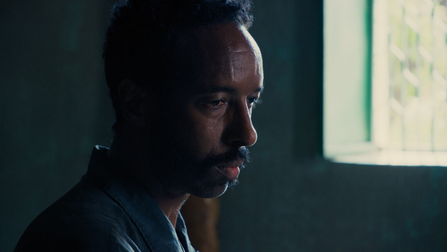 薄暗い室内で、窓から差し込む光を受けながら静かに伏し目がちに佇む男性の横顔の画像。