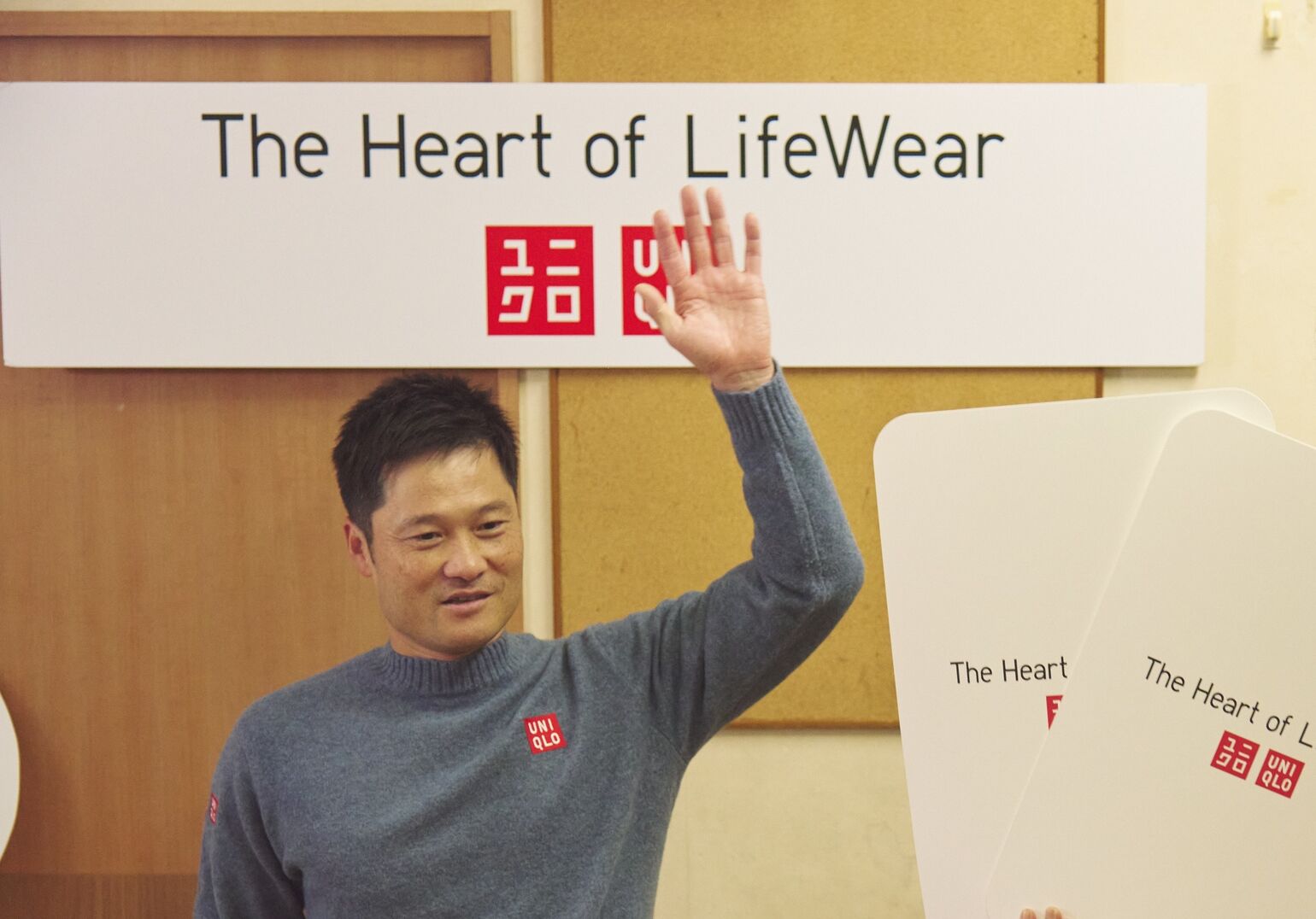 ユニクロのロゴと活動名が入ったボードの前で、青いセーターを着た男性が笑顔で片手を挙げている画像