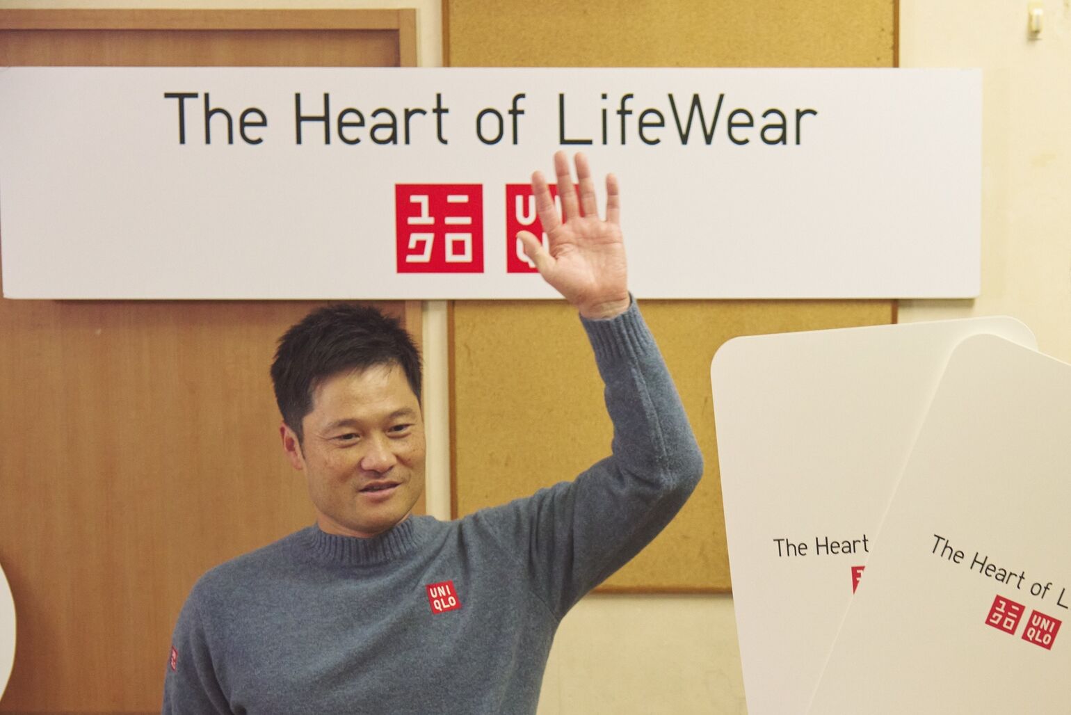 ユニクロのロゴと活動名が入ったボードの前で、青いセーターを着た男性が笑顔で片手を挙げている画像