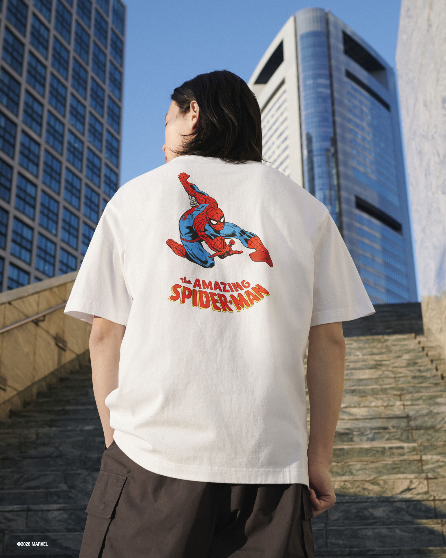 背中に「スパイダーマン」が大きくプリントされた白いTシャツを着たAyumu Matsuokaさんが、高層ビルを背景に階段に立っている後ろ姿の画像。