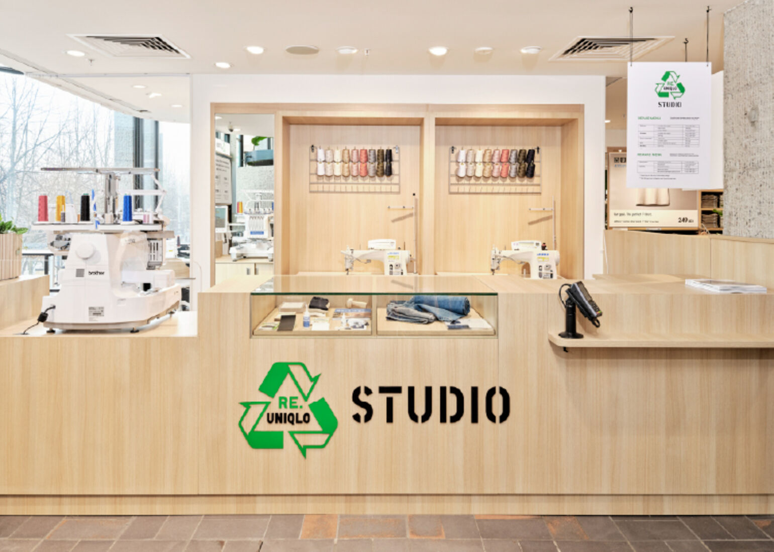 木目調のカウンターに「RE.UNIQLO STUDIO」の緑のリサイクルマークとロゴが描かれた、ユニクロのリペア・リメイク専門ブースの画像。店内には工業用ミシンや多色の糸がディスプレイされており、奥の窓からは外の景色が見える。