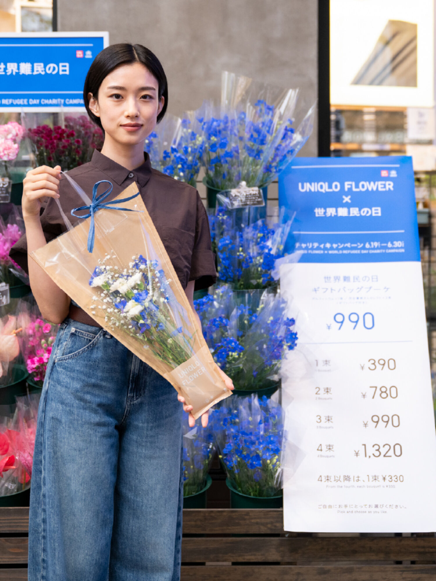 UNIQLO FLOWERの売り場の前で、「世界難民の日ギフトバッグブーケ」を手に持って微笑む俳優の河合優実さん。隣にはキャンペーンの価格表やポスターが設置されている。