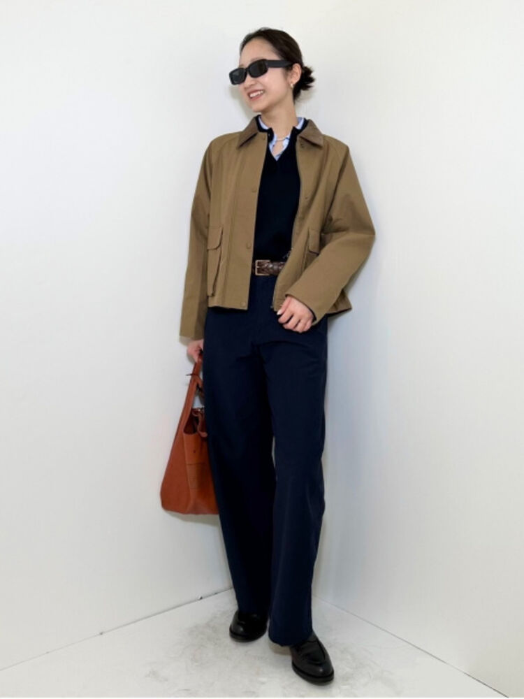 An image of a woman in a traditional full outfit, wearing square-shaped black sunglasses, a brown hunting jacket, and navy pants.