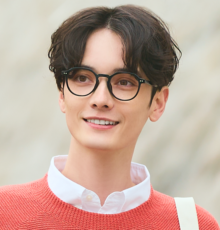 An image of a man wearing classic polygonal frame glasses and an orange knit sweater, looking diagonally forward in a bright outdoor setting.