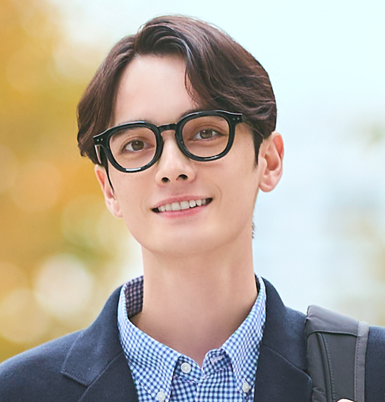 An image of a man wearing thick black-rimmed glasses, paired with a checked shirt and a navy jacket, smiling with an intellectual air.