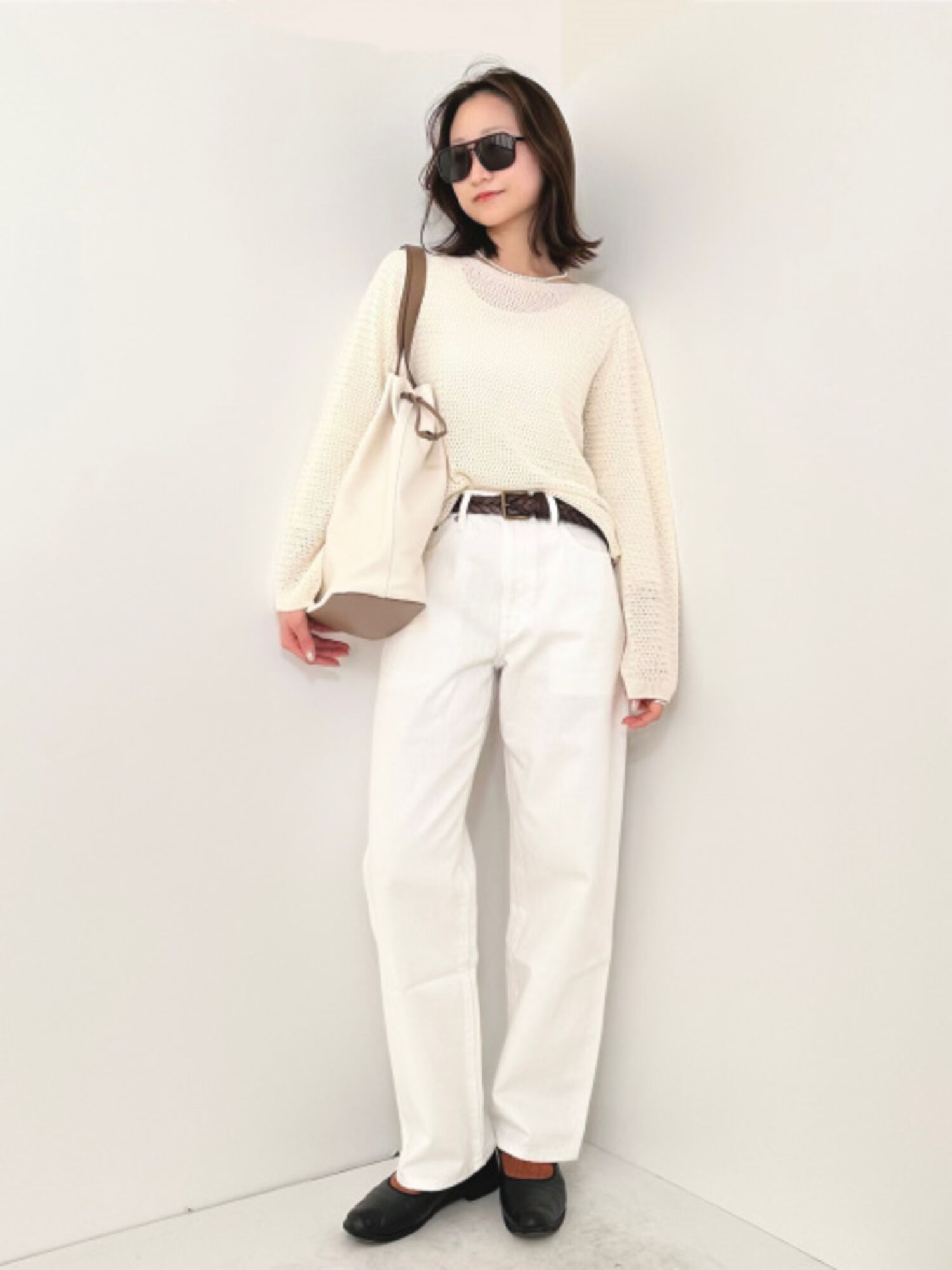 An image of a woman in a full one-tone white outfit, wearing large black teardrop-shaped sunglasses, a white mesh knit, white pants, and a beige bag.