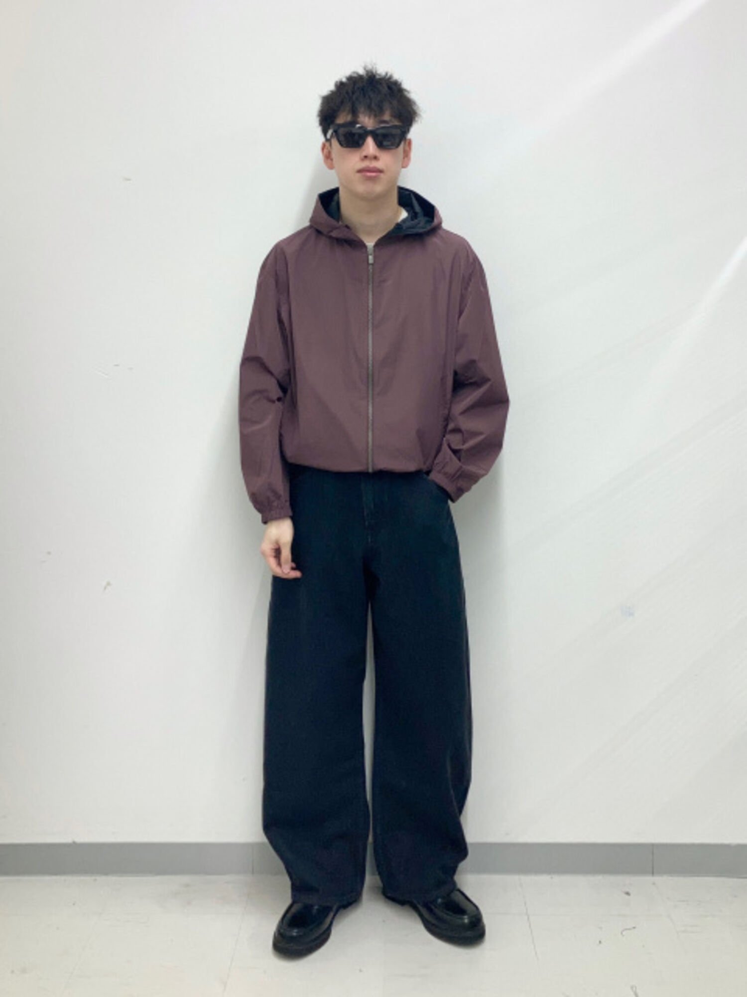 An image of a man in a casual full outfit, wearing standard black-rimmed sunglasses, a brown hoodie jacket, and black wide-leg pants.