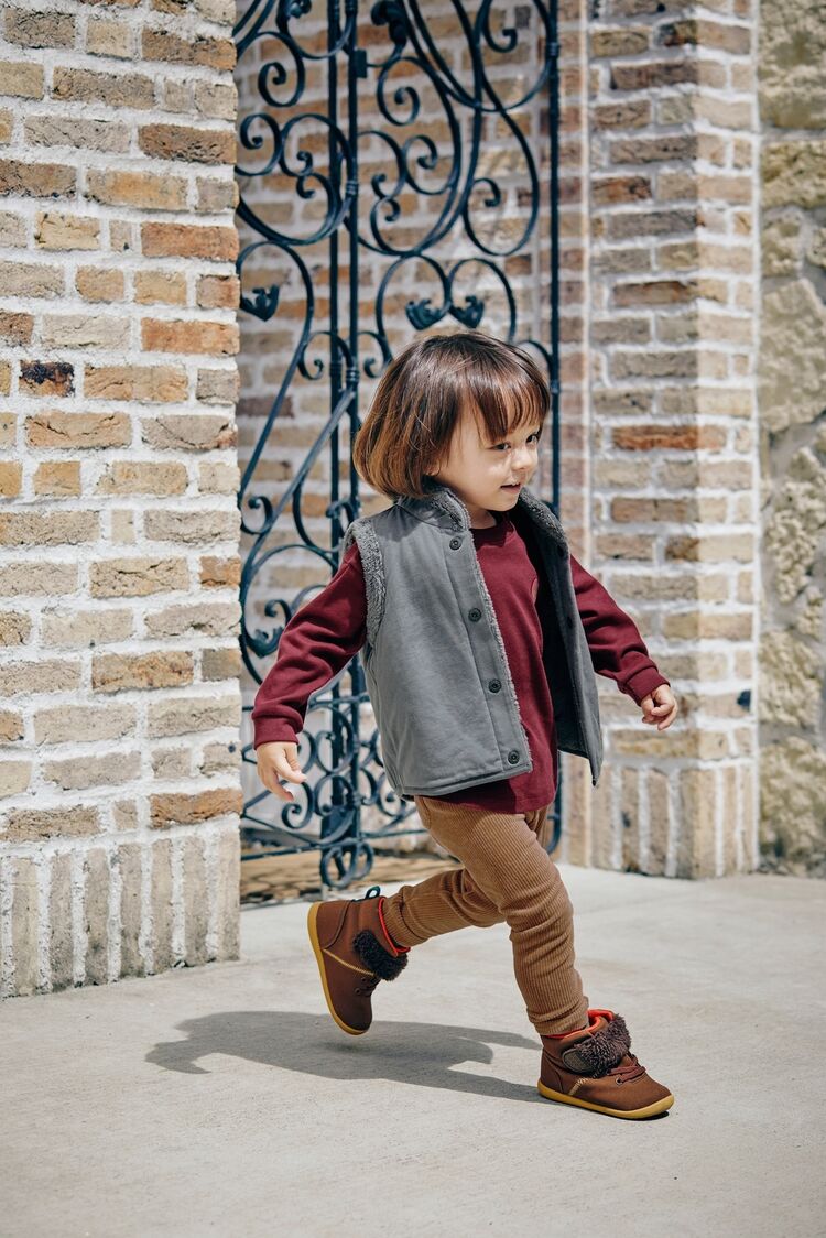 グレーのファーリーフリースリバーシブルベストとワイン色のソフトタッチコットンクルーTを着てブラウンのレギンスを履いた男の子