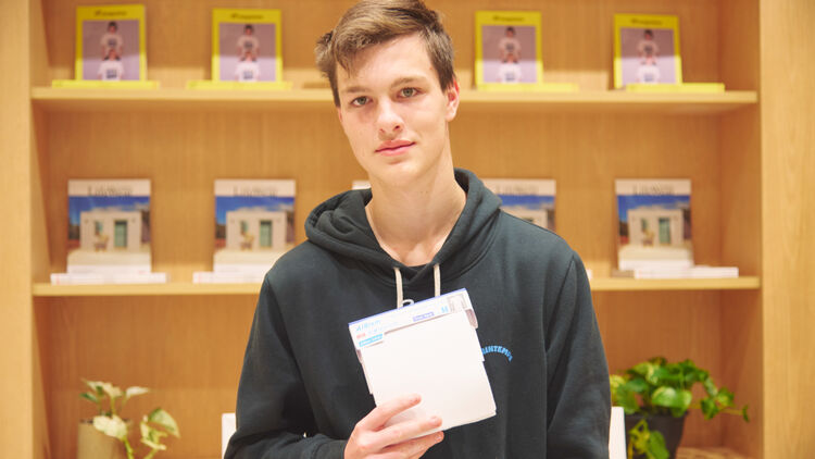 An image of a boy standing in front of a bookshelf, holding a newly received AIRism package carefully with both hands.