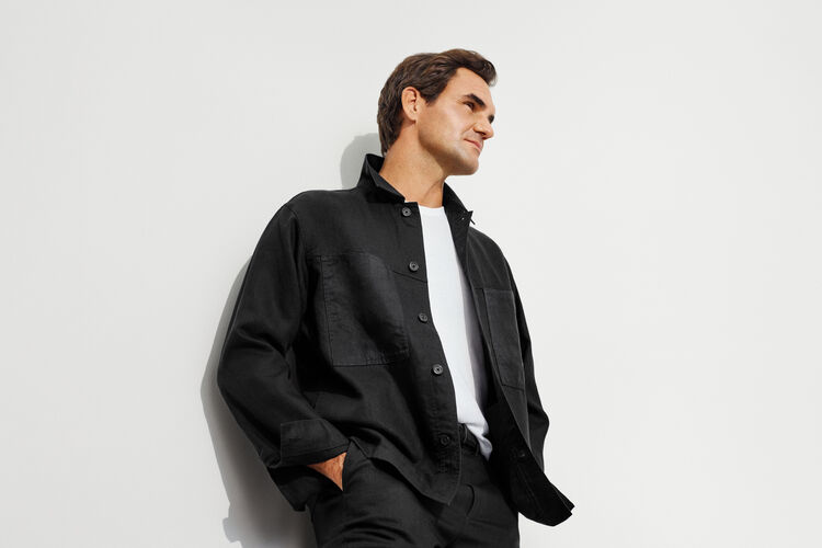 An image of Roger Federer wearing a black linen shirt, leaning against a white wall with a refreshing smile.