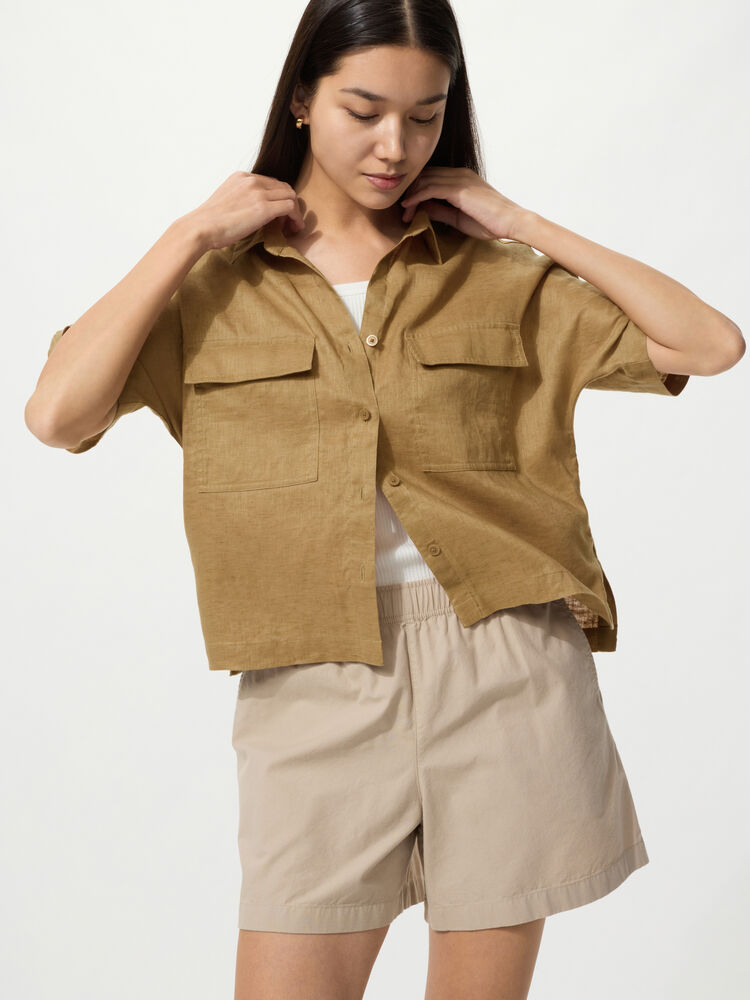 A natural image of a woman wearing an olive-colored linen shirt and beige shorts, looking down with her hand resting on her collar.