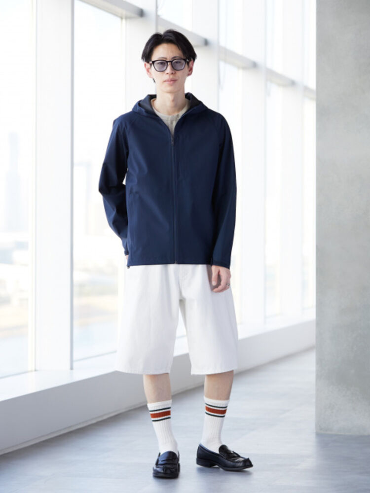 An image of a man in an urban sporty full outfit, wearing black-rimmed glasses, a navy Blocktech parka, white shorts, and striped socks.