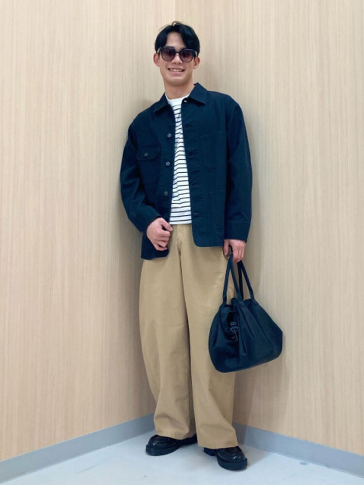 An image of a man in a casual full outfit, wearing slightly tinted square sunglasses, a striped T-shirt, navy coveralls, and beige chino pants.