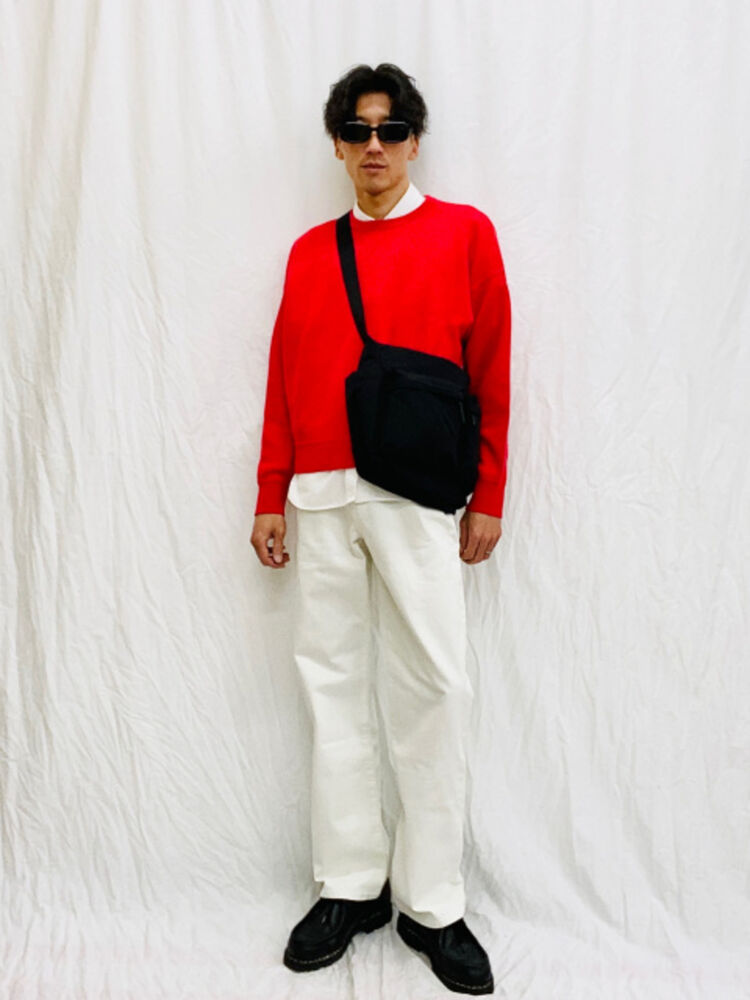 An image of a man in a high-contrast full outfit, wearing narrow-type black sunglasses, a red knit sweater, white pants, and a black shoulder bag.