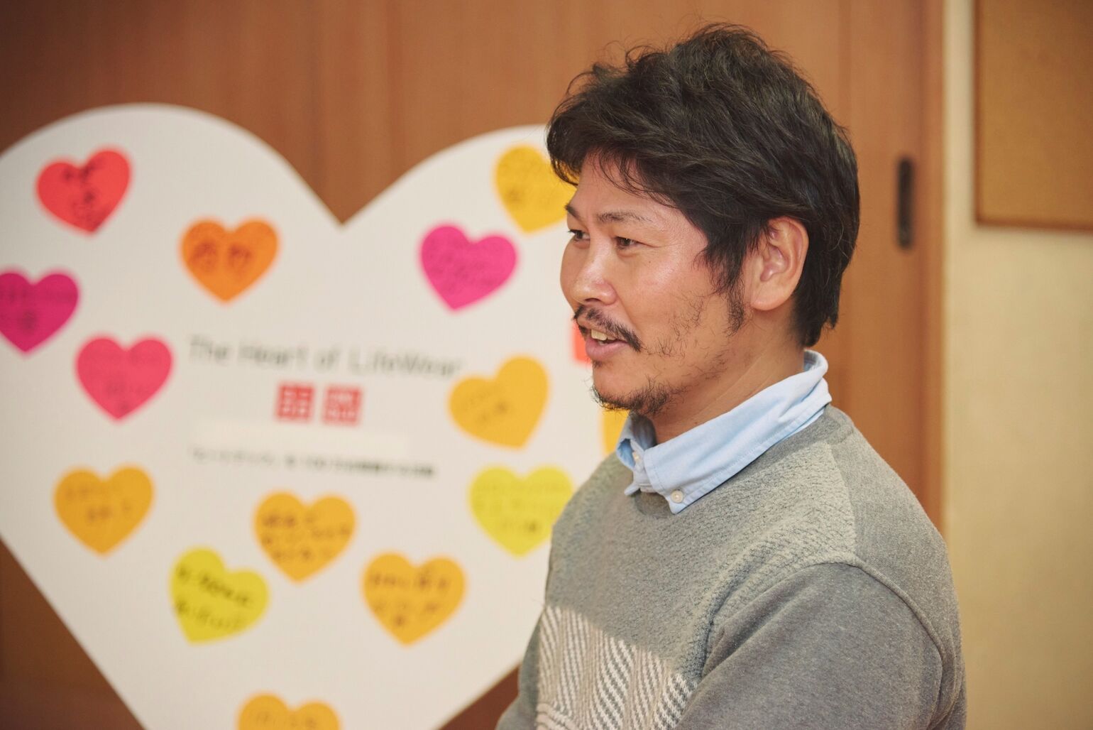 An image of a man looking to the left and speaking, with a heart-shaped board covered in messages in the background.
