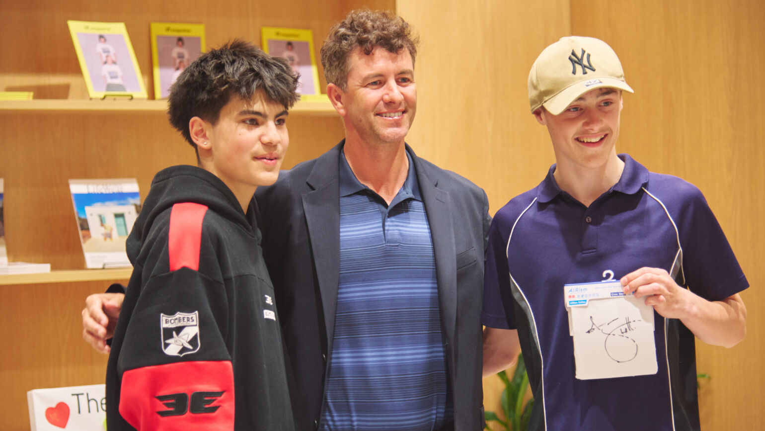 An image of a boy holding a signed AIRism package, with Adam Scott and associates standing next to him with their arms around each other and smiling.