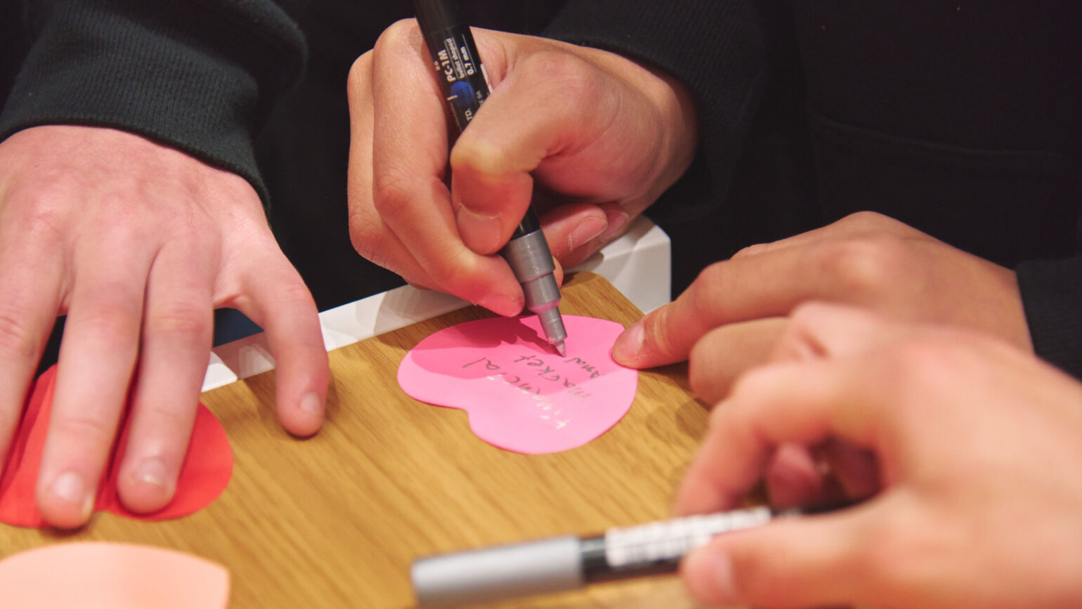 A close-up image of hands using a pen to write a message on a pink heart-shaped card.