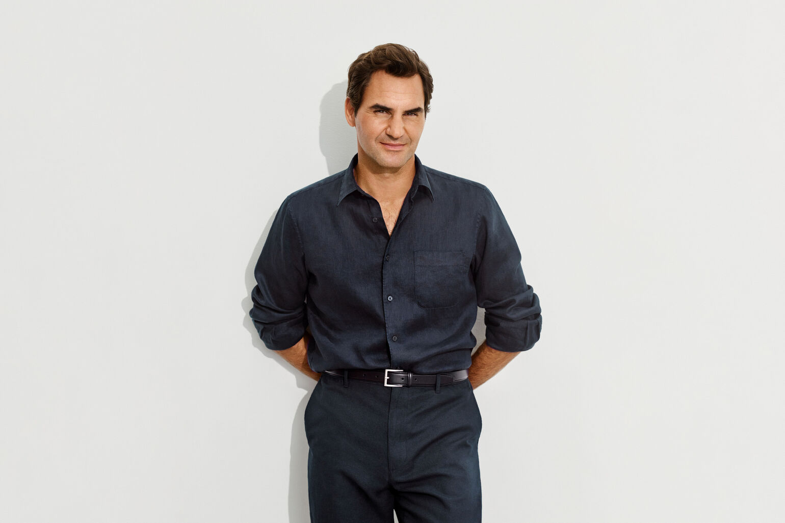 An image of Roger Federer looking smartly dressed in a dark navy linen shirt, standing with his hands on his hips and gazing forward.