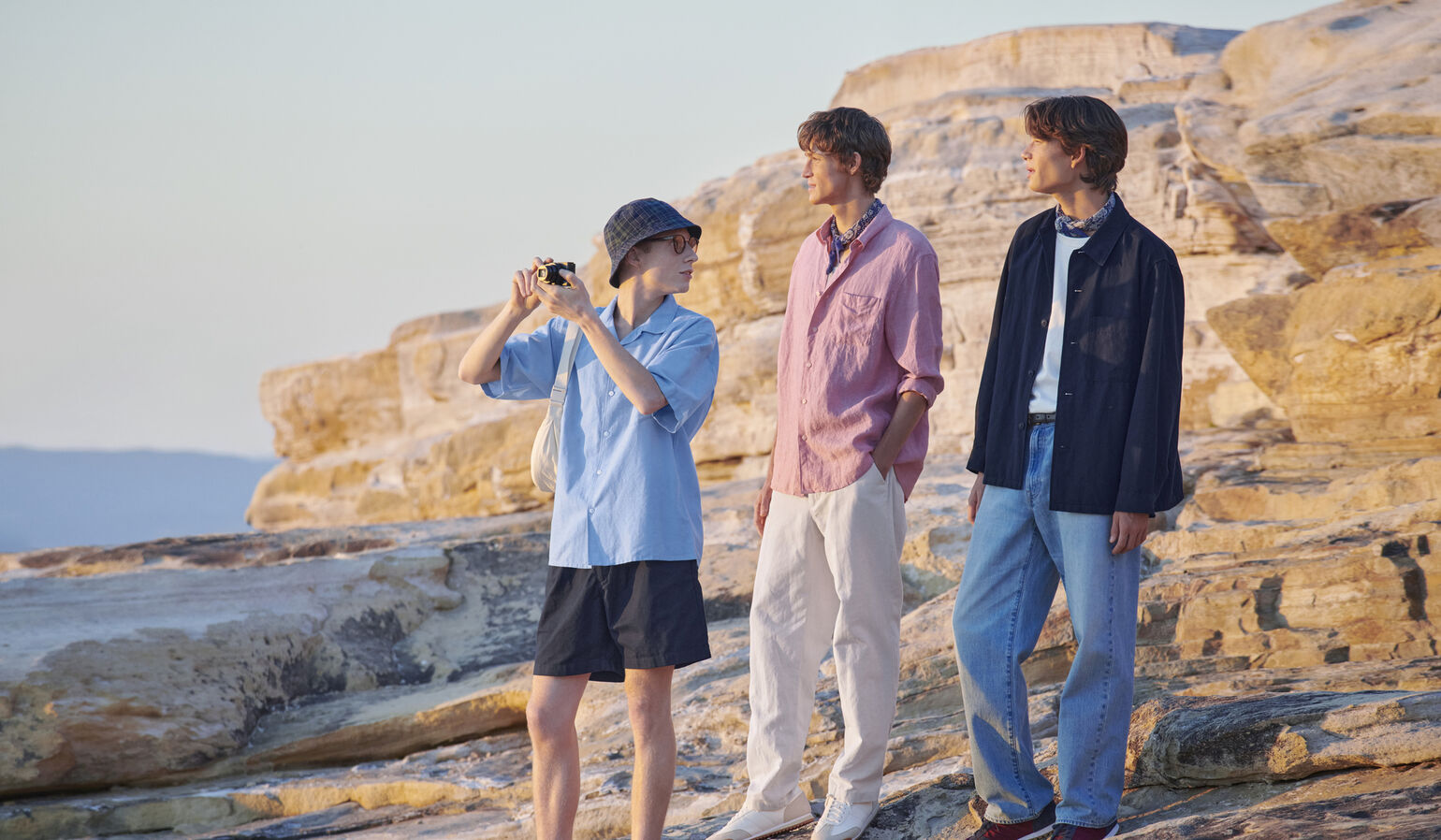 A natural and cheerful image of three men in linen shirts on a rocky shore at sunset, with one of them capturing the scenery with a camera.