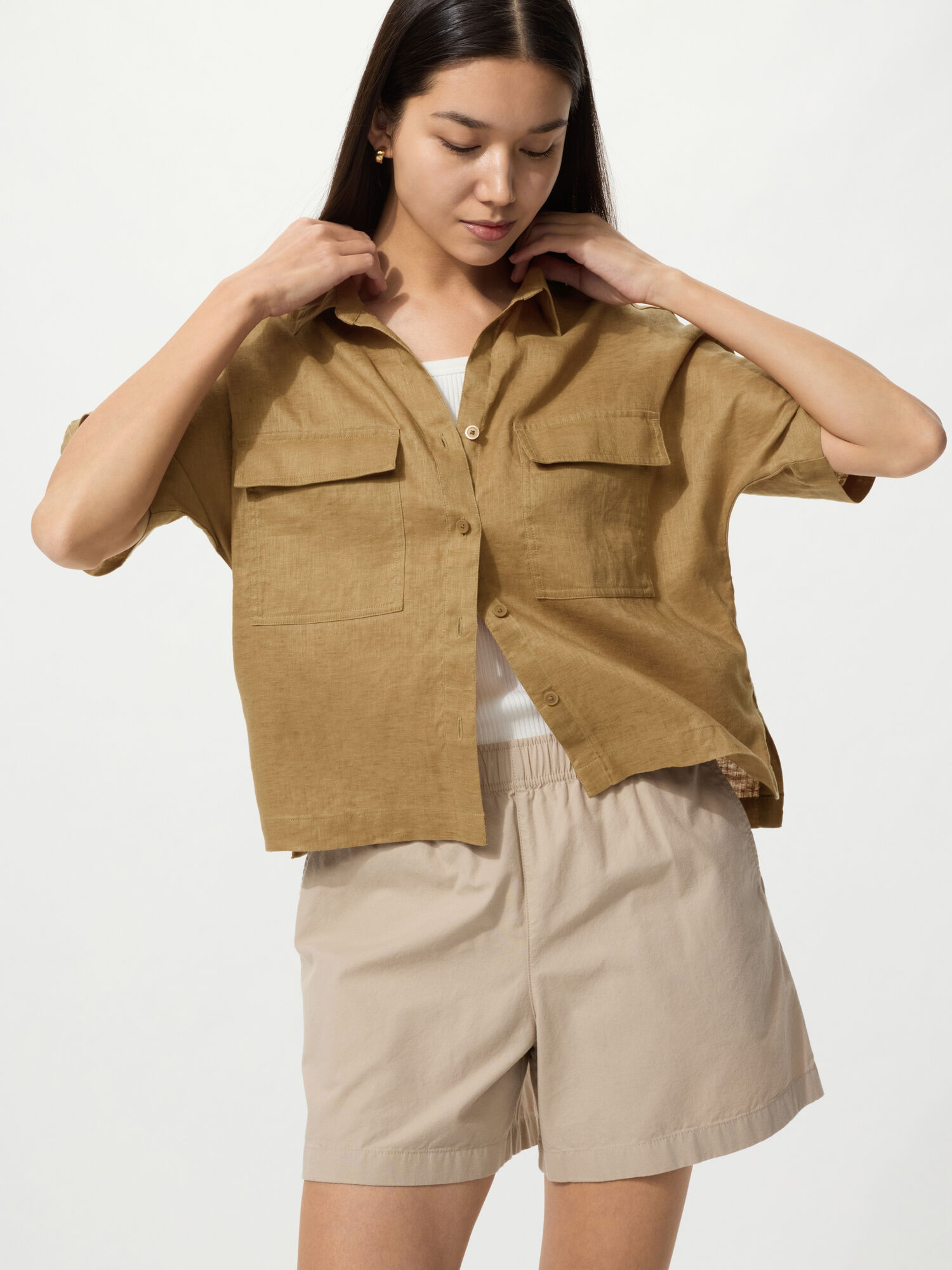 A natural image of a woman wearing an olive-colored linen shirt and beige shorts, looking down with her hand resting on her collar.