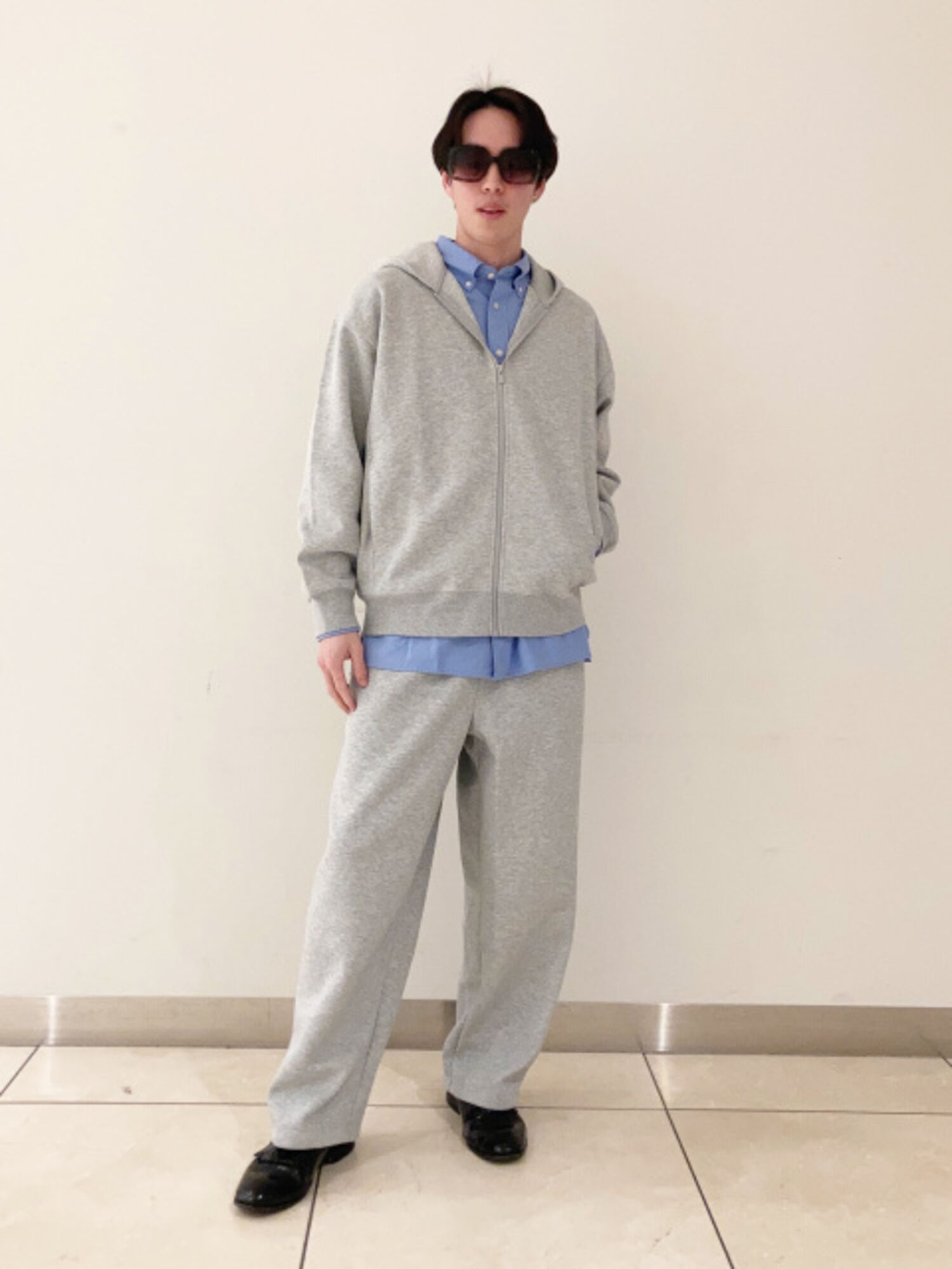 An image of a man in a relaxing full outfit, wearing large square sunglasses, a blue shirt layered over a gray sweatshirt and sweatpants setup.