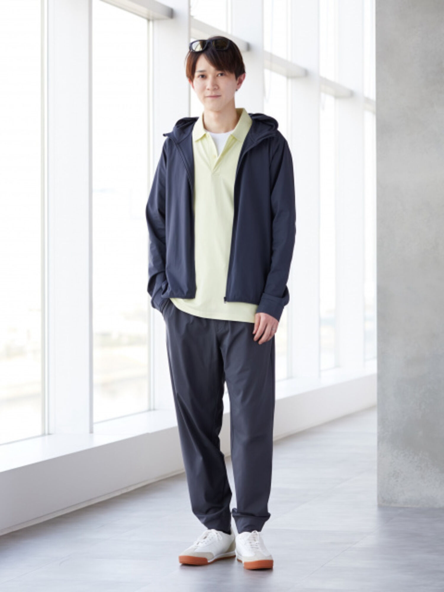 An image of a man in a refreshing full outfit, with sunglasses pushed up onto his head, wearing a pale yellow polo shirt paired with a navy hoodie and jogger pants.