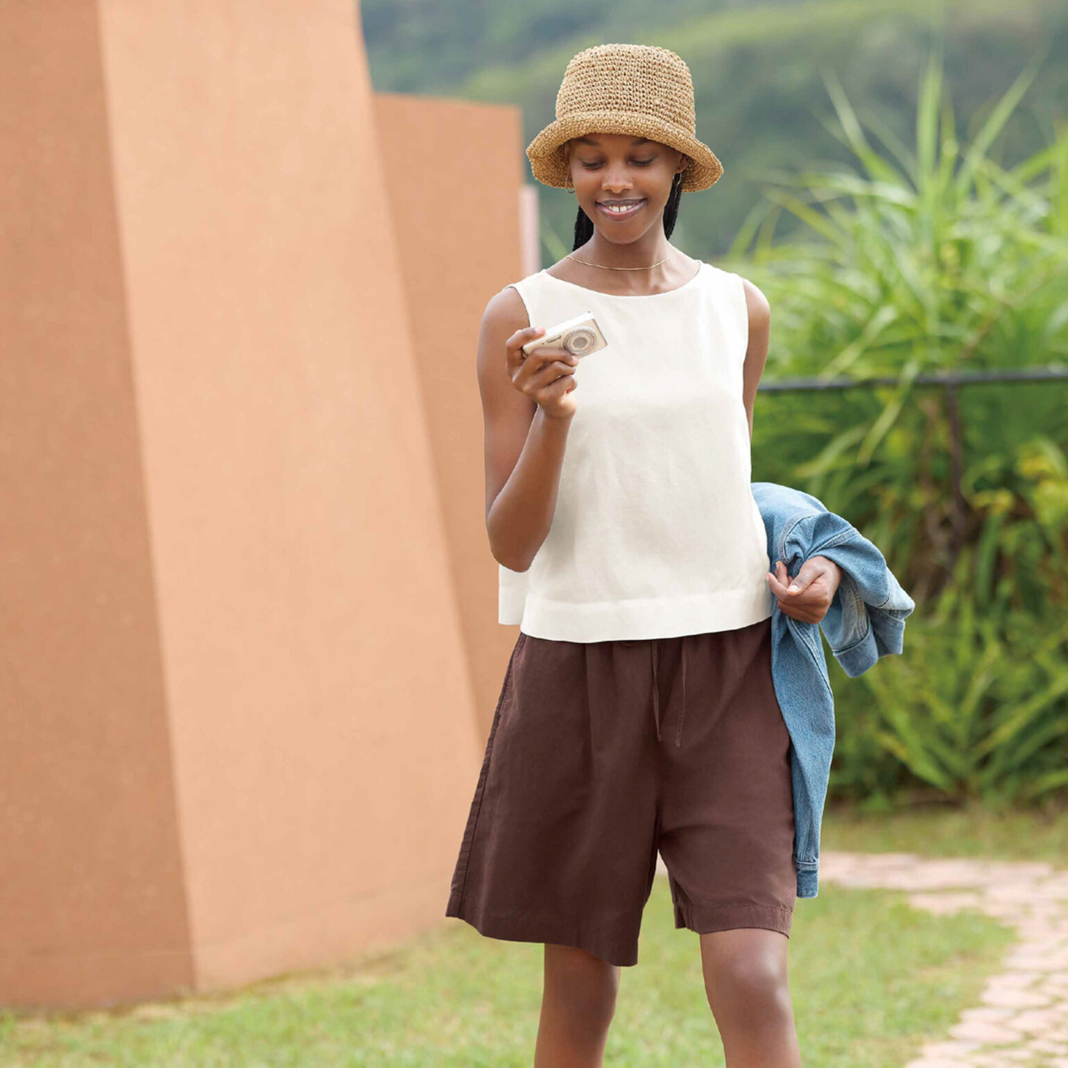 An image of a woman wearing a straw hat, a white sleeveless top, and brown shorts, smiling while looking at a small camera in a lush outdoor setting.