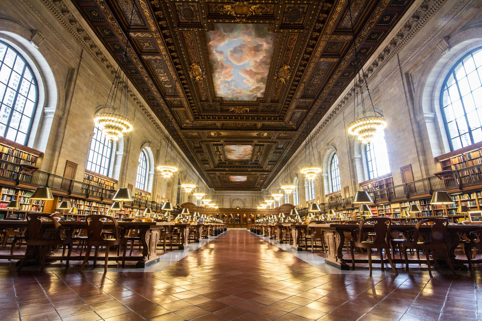 Interior snapshot of the center General Research Division room in the New York Public Library. Brightly lit chandelier lamps, brown carved ceiling with clouded sky painting and rows of wooden seating sided with bookshelves.