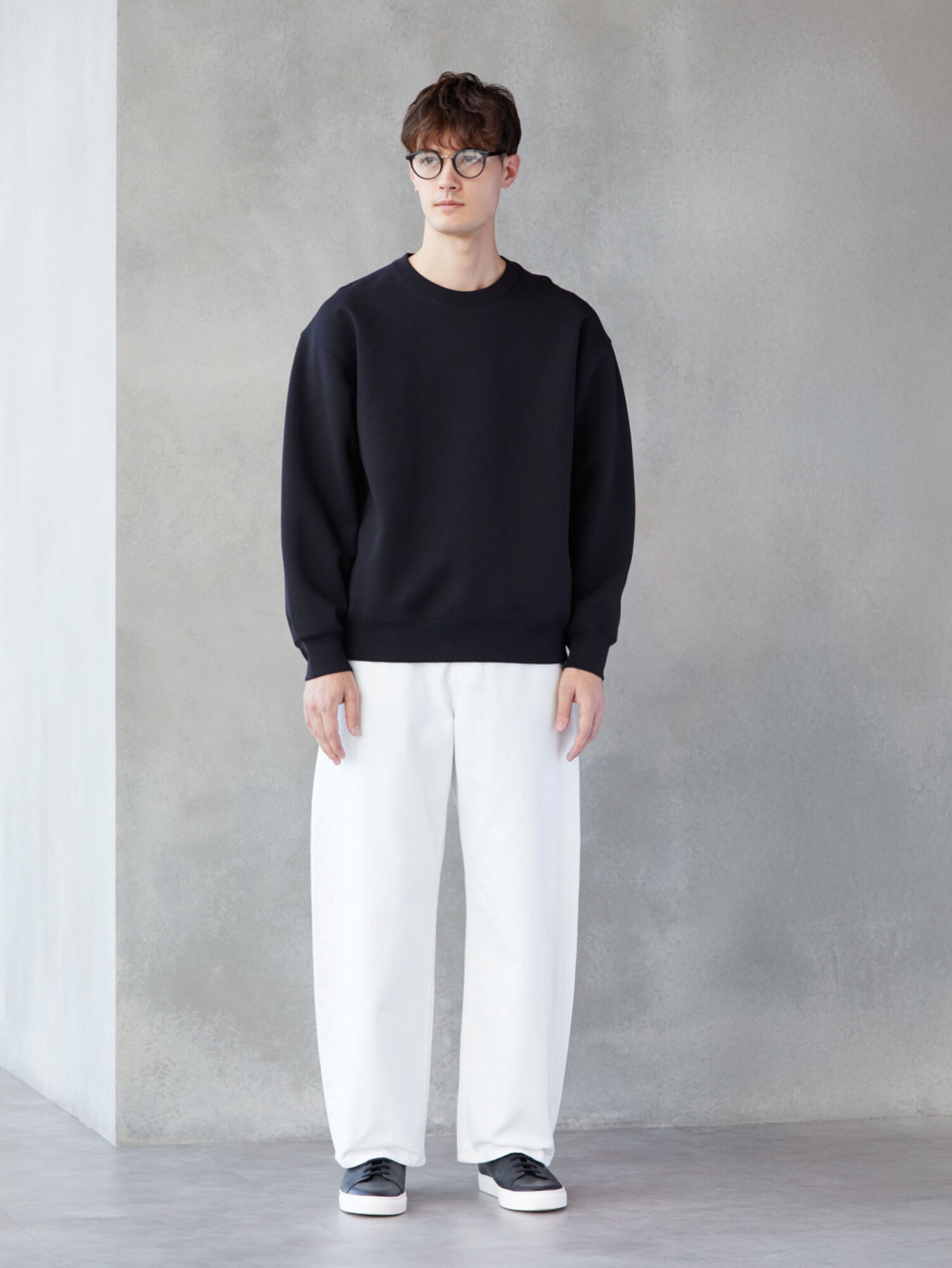 An image of a man in a clean, monotone full outfit, wearing black-rimmed glasses, a simple black sweatshirt, and white wide-leg pants.