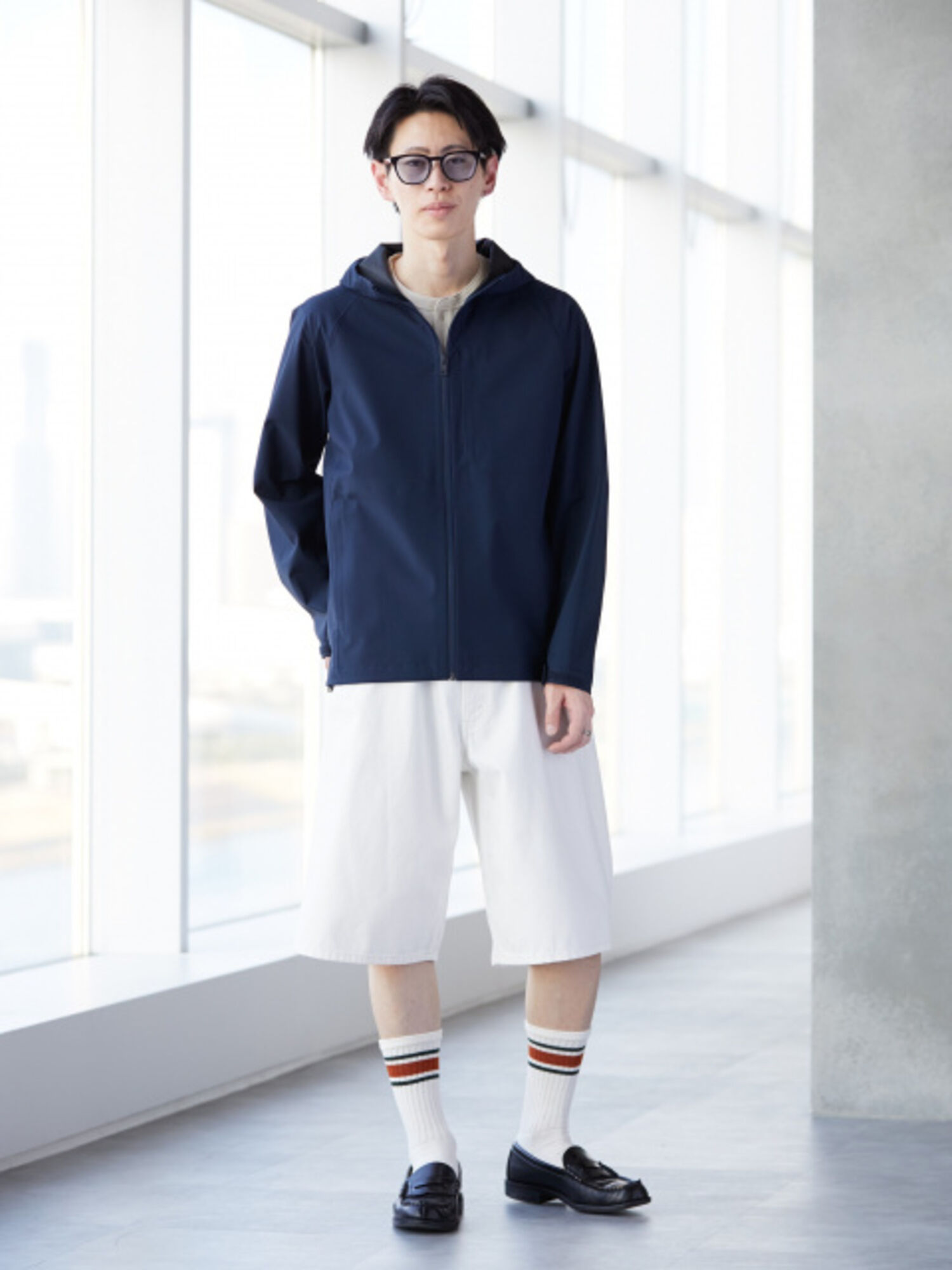An image of a man in an urban sporty full outfit, wearing black-rimmed glasses, a navy Blocktech parka, white shorts, and striped socks.