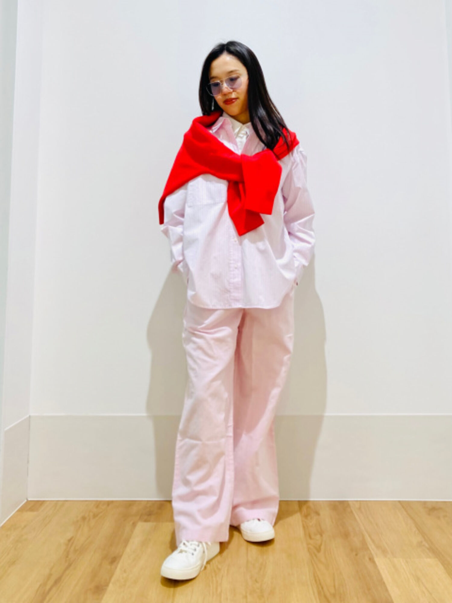 An image of a woman in a glamorous full outfit, wearing light-colored lens sunglasses, a pink striped shirt, and a red knit sweater draped over her shoulders.